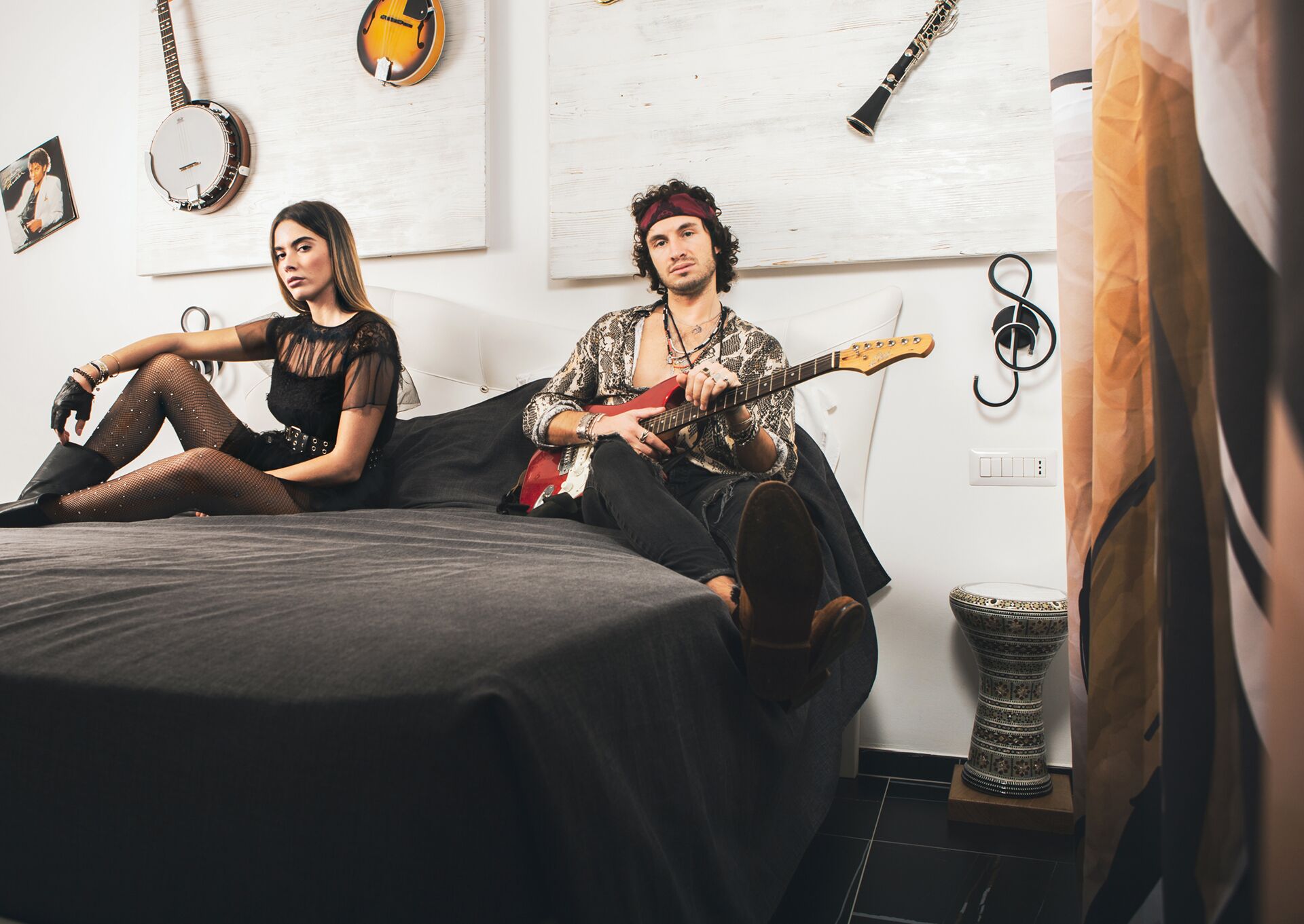 Two people sitting on a bed in a room with musical instruments on the wall.