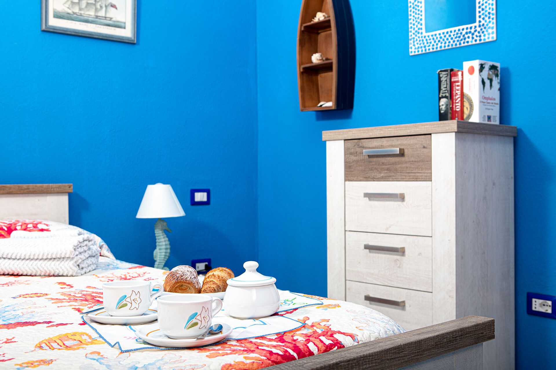 A bedroom with a bed, a bedside table with a lamp, a dresser, and a wall with a framed picture and a shelf with books.