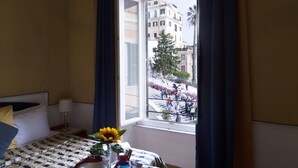 Ein Hotelzimmer mit Bett, Nachttisch, Fenster mit Vorhängen und Blick auf eine belebte Straße mit Menschen und Gebäuden.