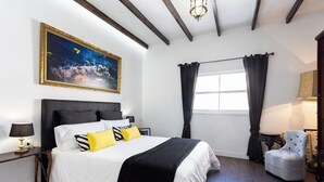 Un dormitorio con una cama grande, una mesita de noche, una silla, una ventana con cortinas y un cuadro en la pared.