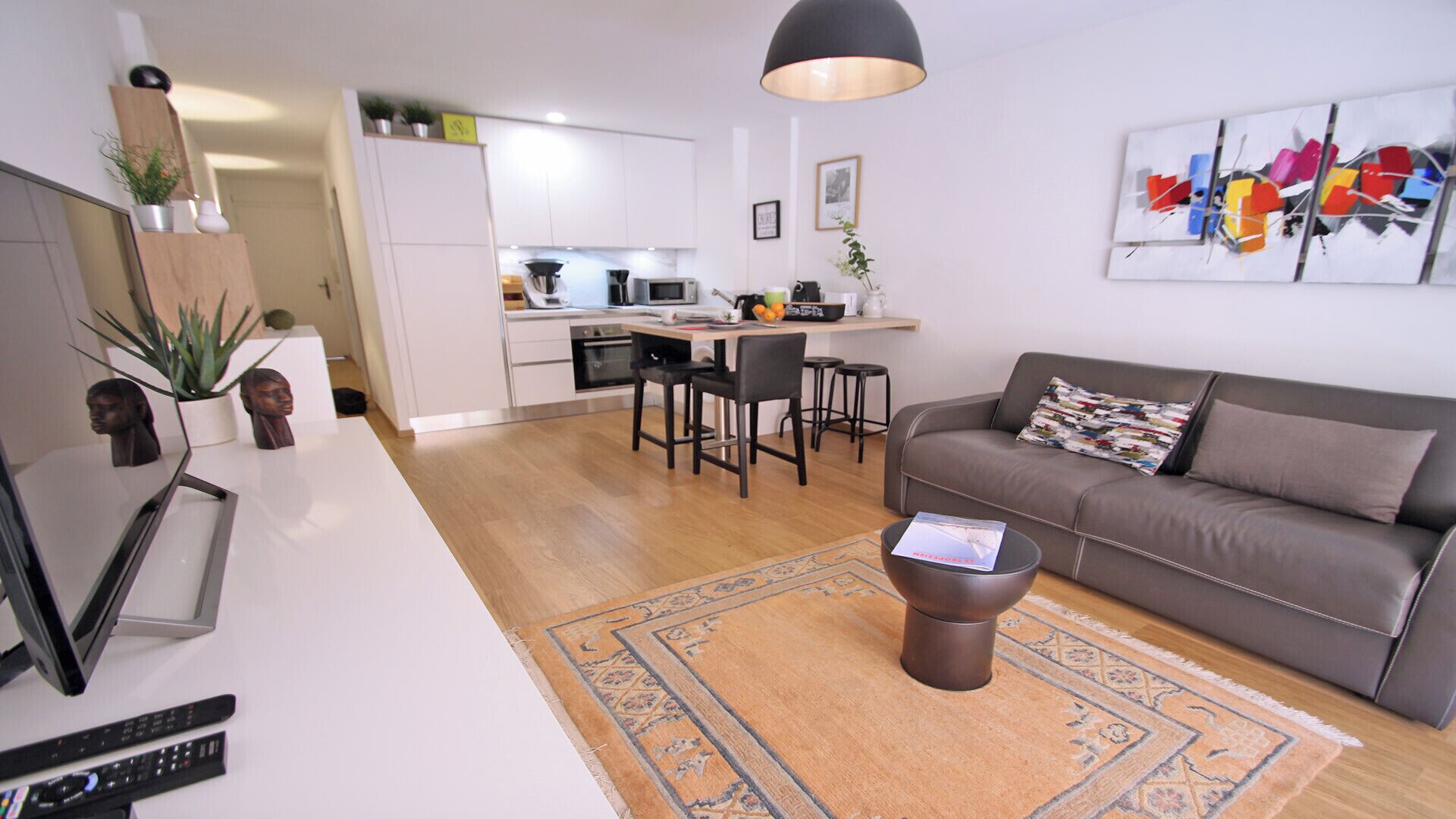 Ein modernes Wohnzimmer mit einer Couch, einem Couchtisch und einem Teppich. Es gibt eine Küchenzeile mit weißen Schränken und einem Esstisch.