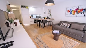 Una sala de estar moderna con sofá, mesa de centro y alfombra. Hay una zona de cocina con armarios blancos y una mesa de comedor.