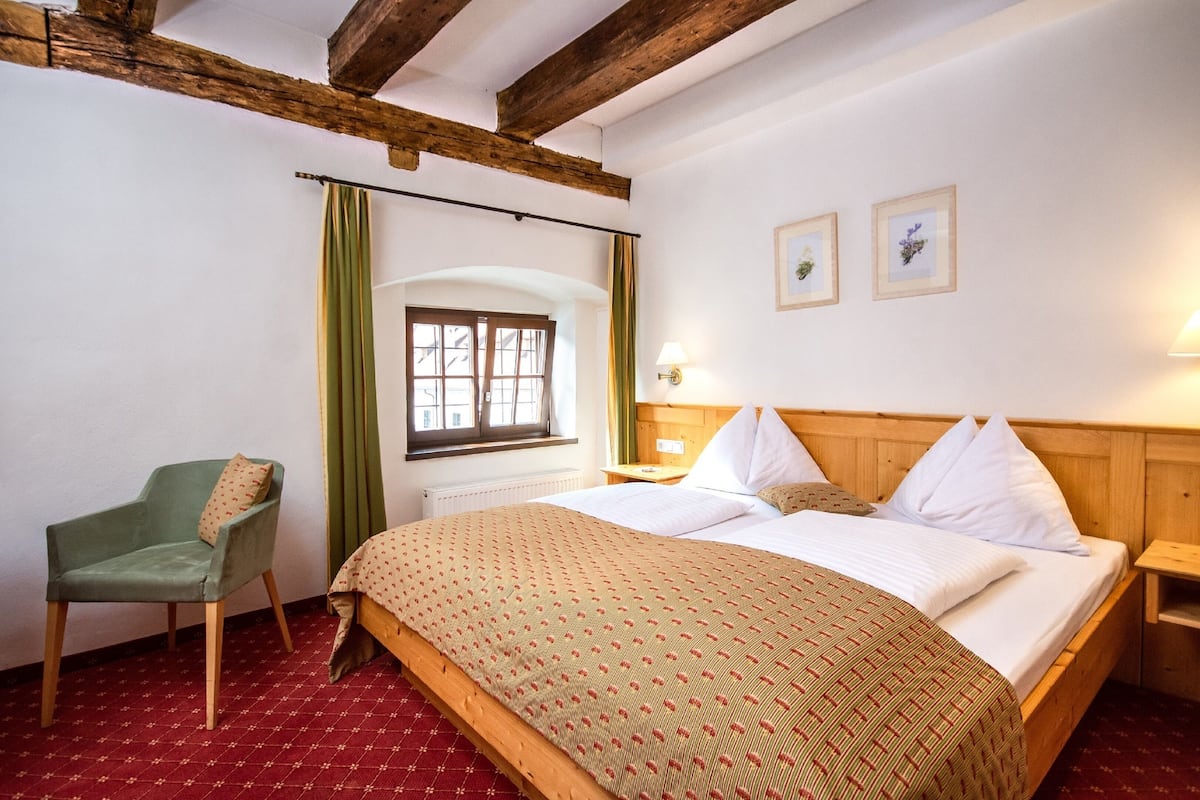 A room with a wooden bed, a green chair, a window with curtains, and two framed pictures on the wall.