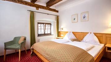 A room with a wooden bed, a green chair, a window with curtains, and two framed pictures on the wall.