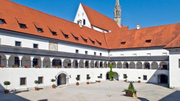 A courtyard with white buildings, red roofs, and a church spire in the background. - Hotel Kartause (Gaming)