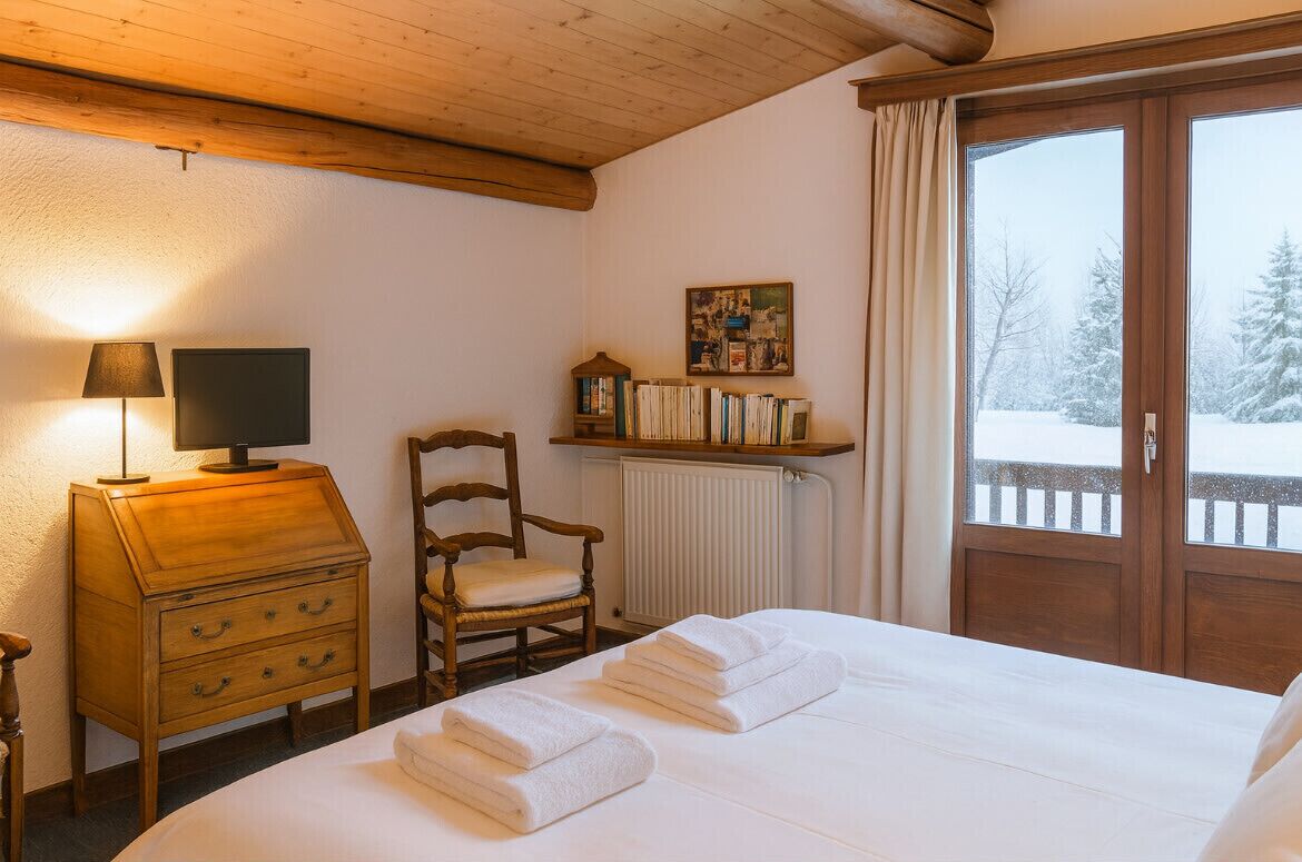 A cozy hotel room with a wooden bed, a desk with a lamp, a chair, a bookshelf, and a window with a snowy view.