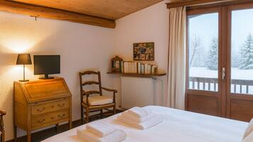 A cozy hotel room with a wooden bed, a desk with a lamp, a chair, a bookshelf, and a window with a snowy view.
