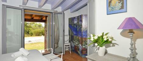 Une chambre à coucher avec un lit, une chaise, une table, une lampe, un vase avec des fleurs et un tableau au mur.