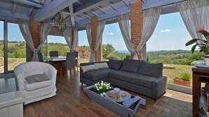 Una sala de estar moderna con un sofá gris, un sillón blanco y una zona de comedor con mesa y sillas. Amplias ventanas ofrecen vistas al campo.