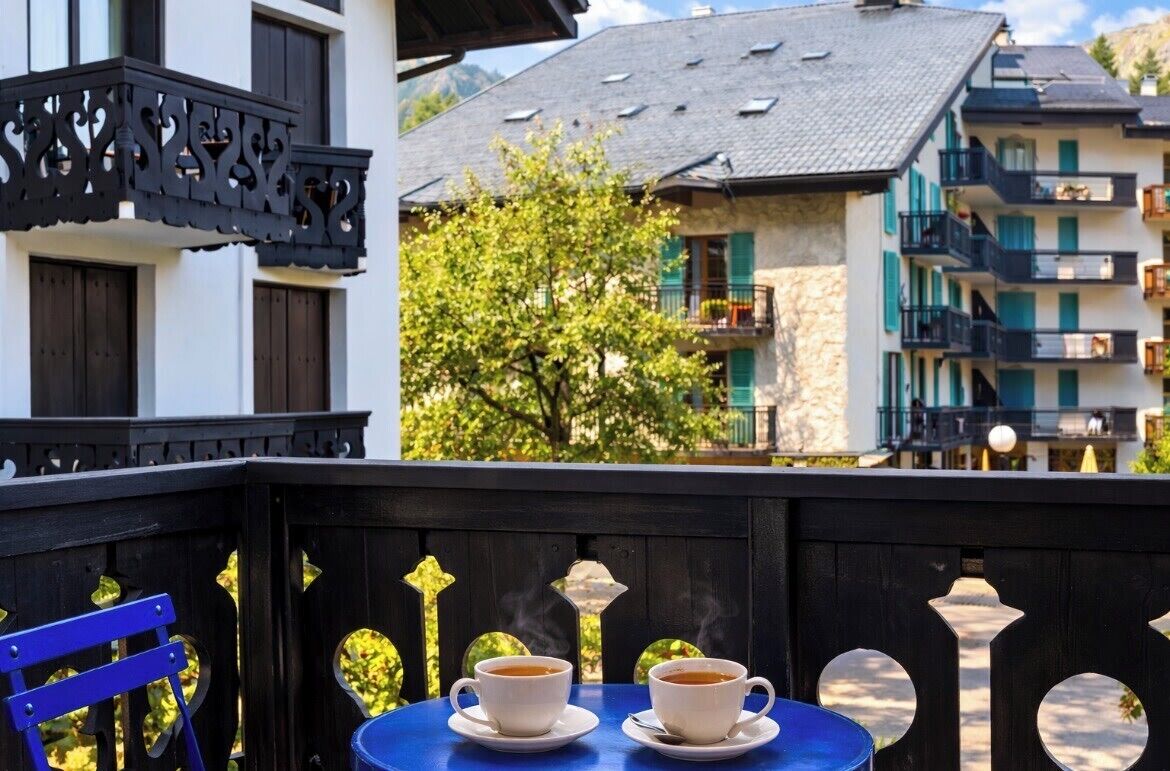 Ein Balkon mit zwei weißen Kaffeetassen, einem blauen Stuhl und Blick auf ein Gebäude mit Balkonen und einem Baum.