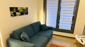 A living room with a blue sofa, a patterned rug, a painting on the wall, and a window with blinds. - Cosy Nest Near Center Chamonix (Chamonix-Mont-Blanc)