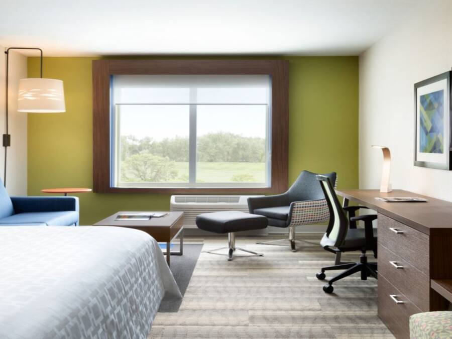 A hotel room with a bed, desk, chair, and a large window offering a view of greenery.