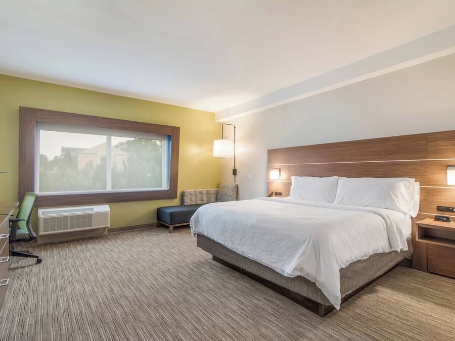A hotel room with a large bed, bedside tables, a desk with a chair, a window, and a wall-mounted lamp.