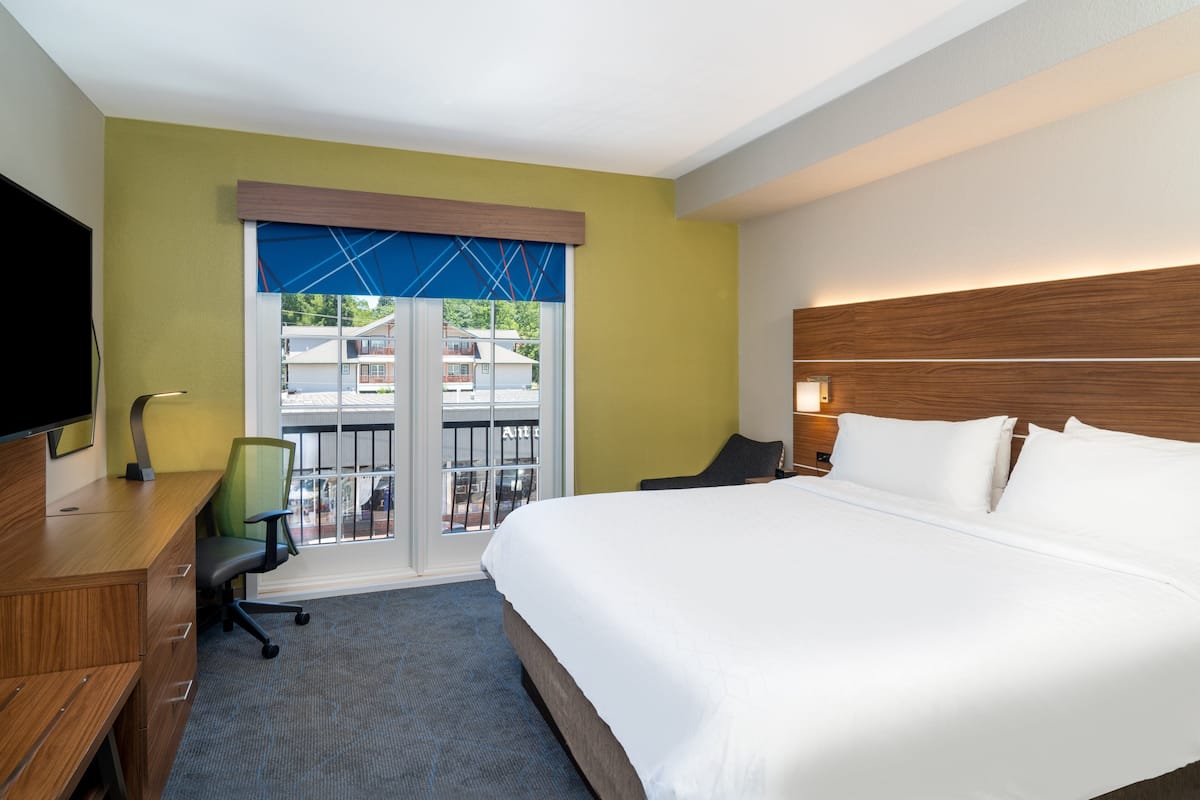 A hotel room with a large bed, a desk with a chair, a TV, and a window with a view of buildings outside.