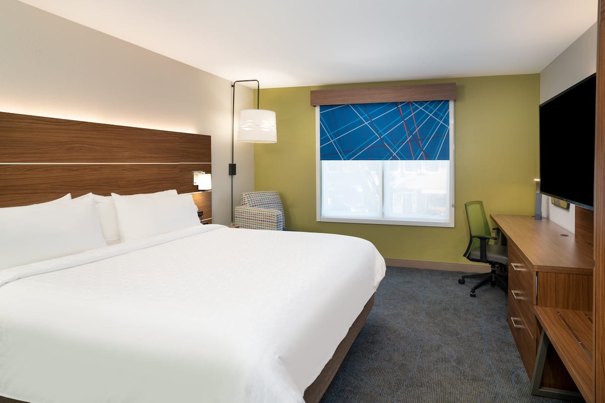 A hotel room with a large bed, a desk with a chair, a TV, and a window with a geometric view.