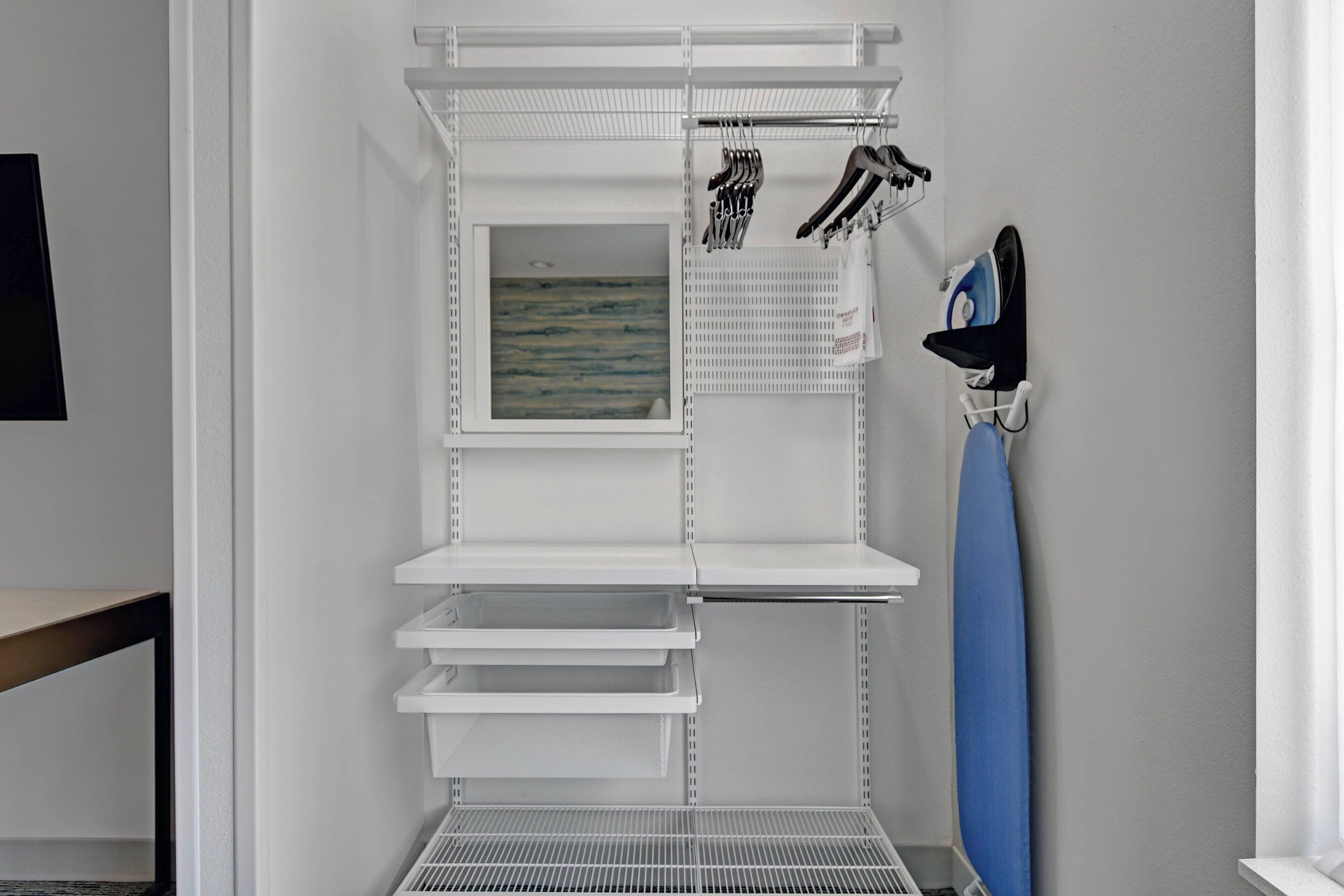 A white metal shelving unit with multiple compartments, a small framed artwork, and a blue ironing board.