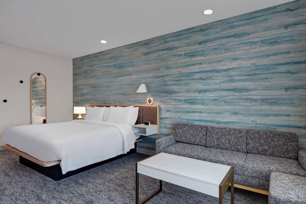 A modern hotel room with a large bed, a grey sofa, a white coffee table, and a wall with a blue abstract design.