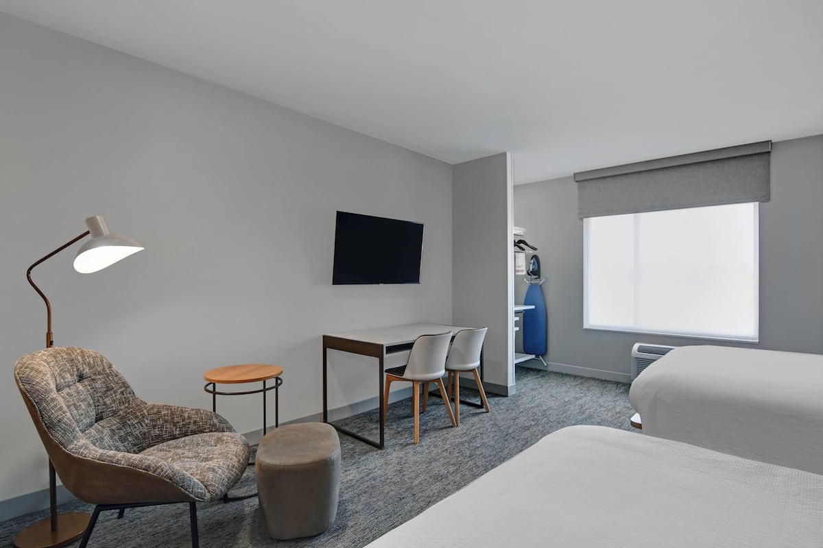 A hotel room with a single bed, a desk with two chairs, a standing lamp, a flat-screen TV, and a large window.