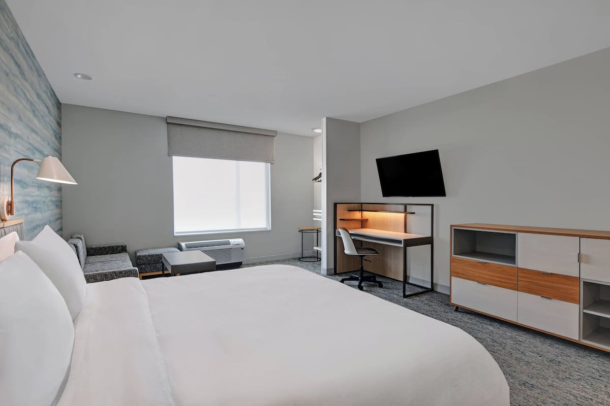 A hotel room with a large bed, a desk with a chair, a TV mounted on the wall, and a window with a view.