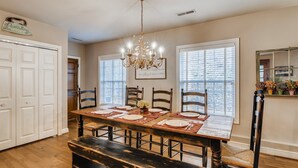 A dining room with a wooden table set for four, surrounded by chairs, a chandelier, and a large window with blinds. - Over Yonder (Leipers Fork)