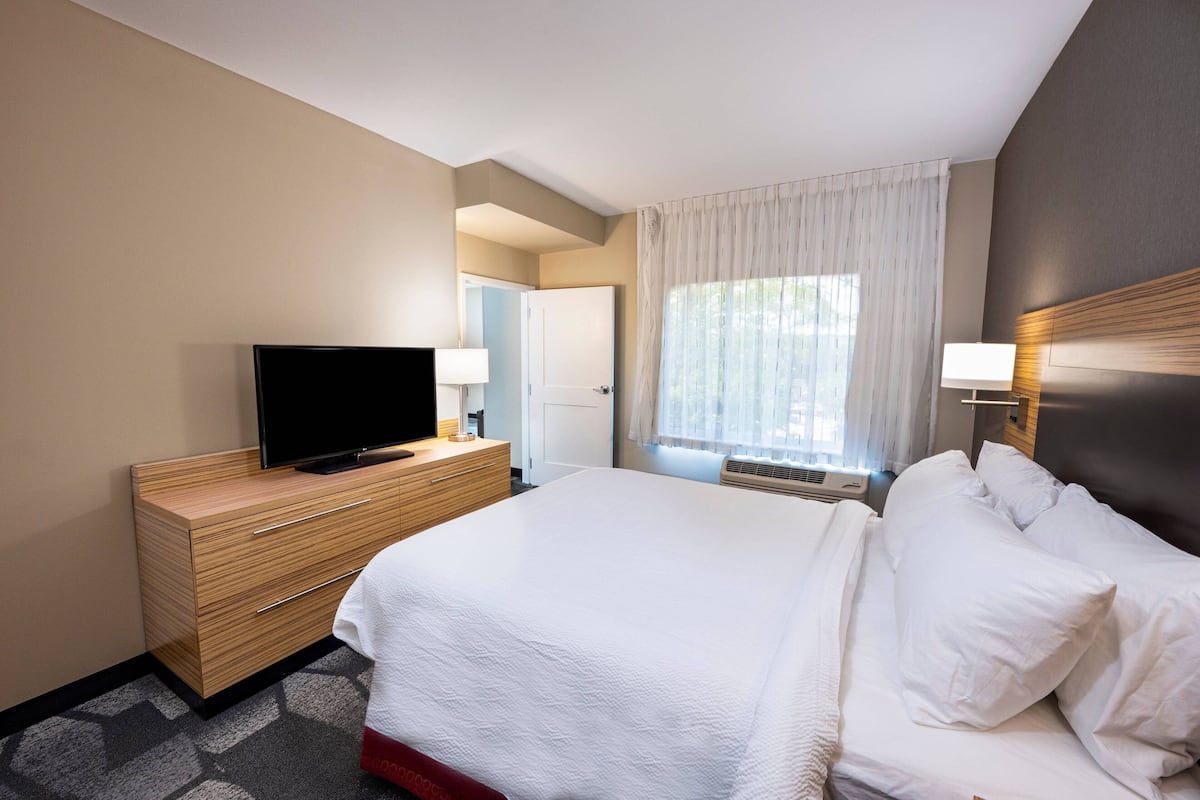 Een hotelkamer met een bed, een televisie op een houten dressoir, een raam met gordijnen en een nachtkastje met een lamp.