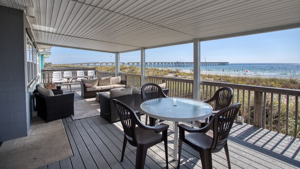 A covered outdoor deck with a round table and chairs, overlooking a beach and pier.