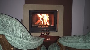 Un rincón acogedor con dos sillas, una mesita y una chimenea encendida. Se observa una copa de vino sobre la mesa.