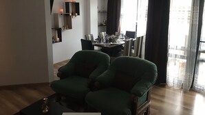 Una sala de estar con dos sillones verdes, una mesa de comedor preparada para cuatro personas y un ventanal con cortinas.