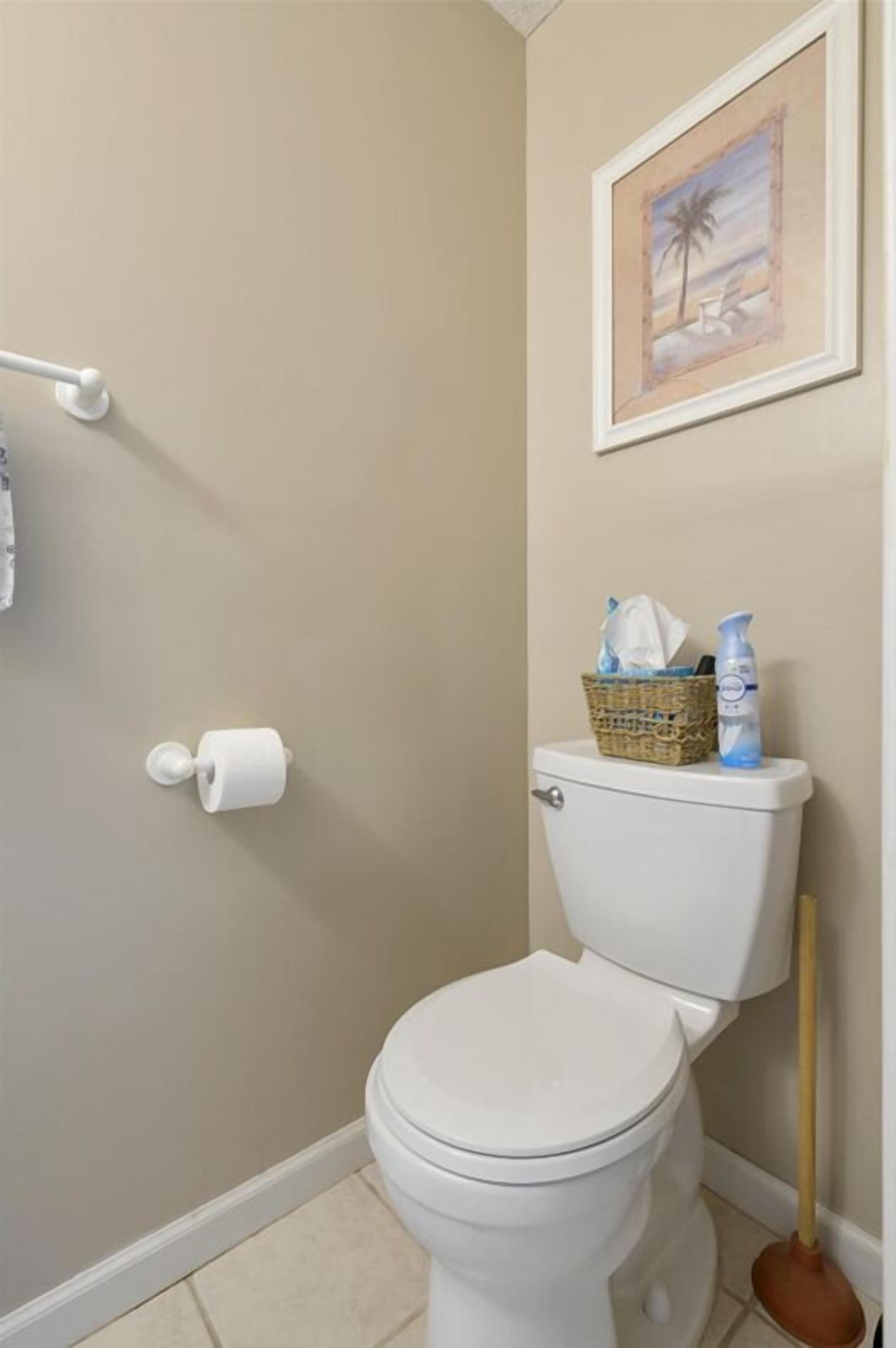 A bathroom with a toilet, a roll of toilet paper, a plunger, and a basket with cleaning supplies on the wall.