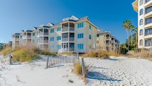 Un hotel frente a la playa con varios balcones, una playa arenosa en primer plano y un cielo azul despejado.