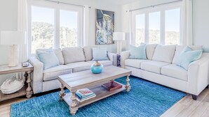 Una sala de estar con un sofá seccional de tonos claros, una mesa de centro de madera y un tapete azul. Se ven dos ventanas con cortinas blancas, y un cuadro en la pared.