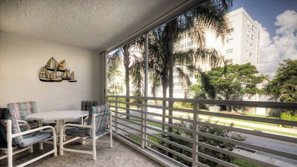 Een balkon met een witte tafel en stoelen, een decoratieve wandversiering en uitzicht op palmbomen en gebouwen.