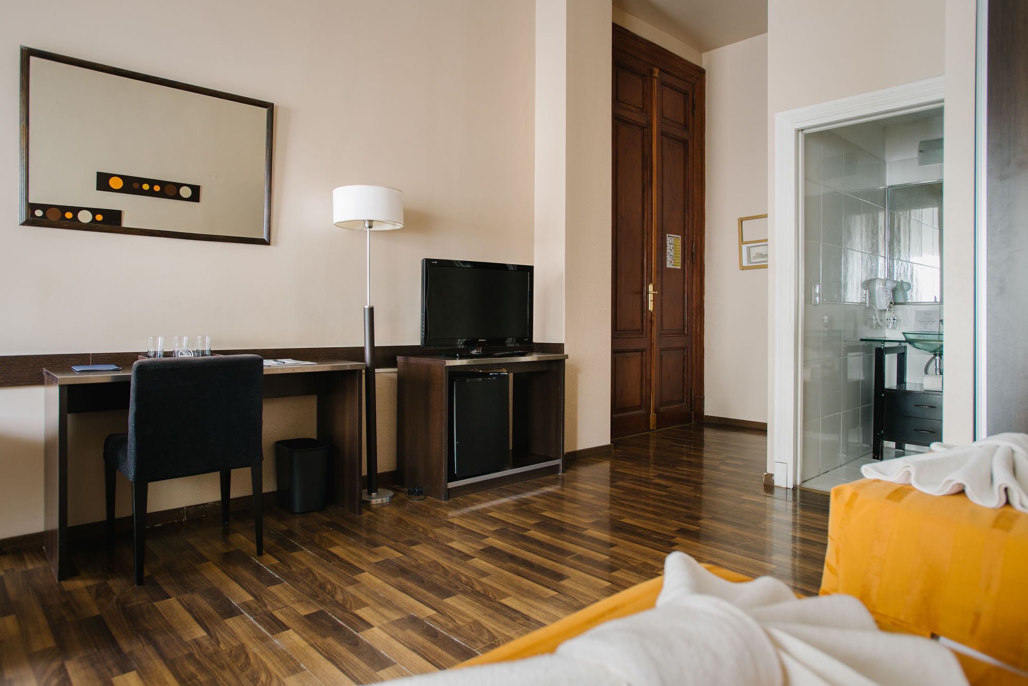 A hotel room with a wooden floor, a desk with a computer and a TV, a bed with a white and yellow blanket, and a bathroom visible through a glass door.
