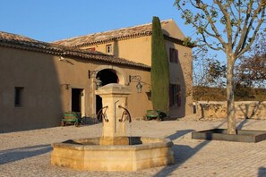 Une cour avec une fontaine, un bâtiment doté d’un toit en tuiles et un arbre.