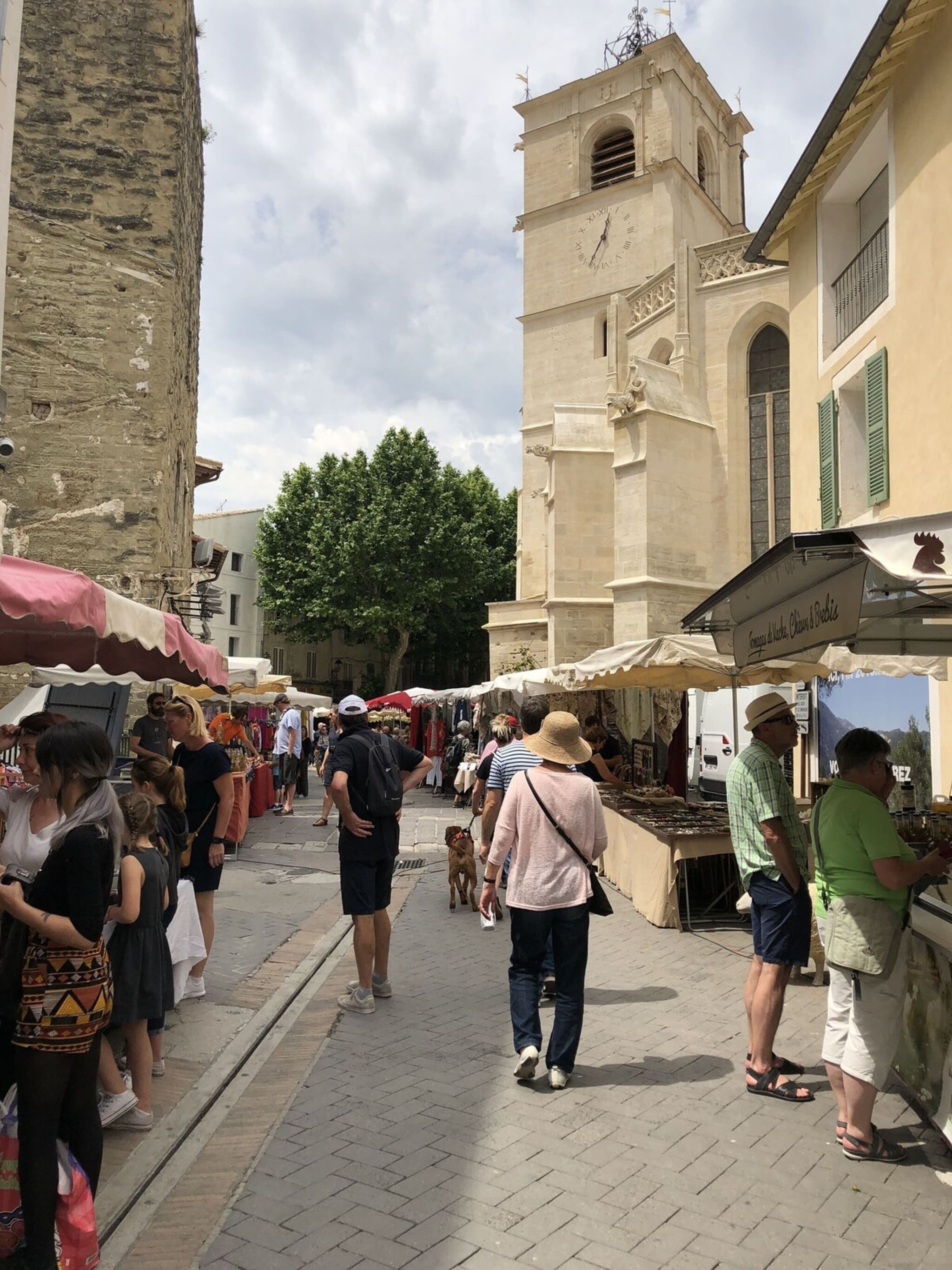 Un mercado al aire libre lleno de gente comprando, con un edificio histórico y una torre de reloj al fondo.