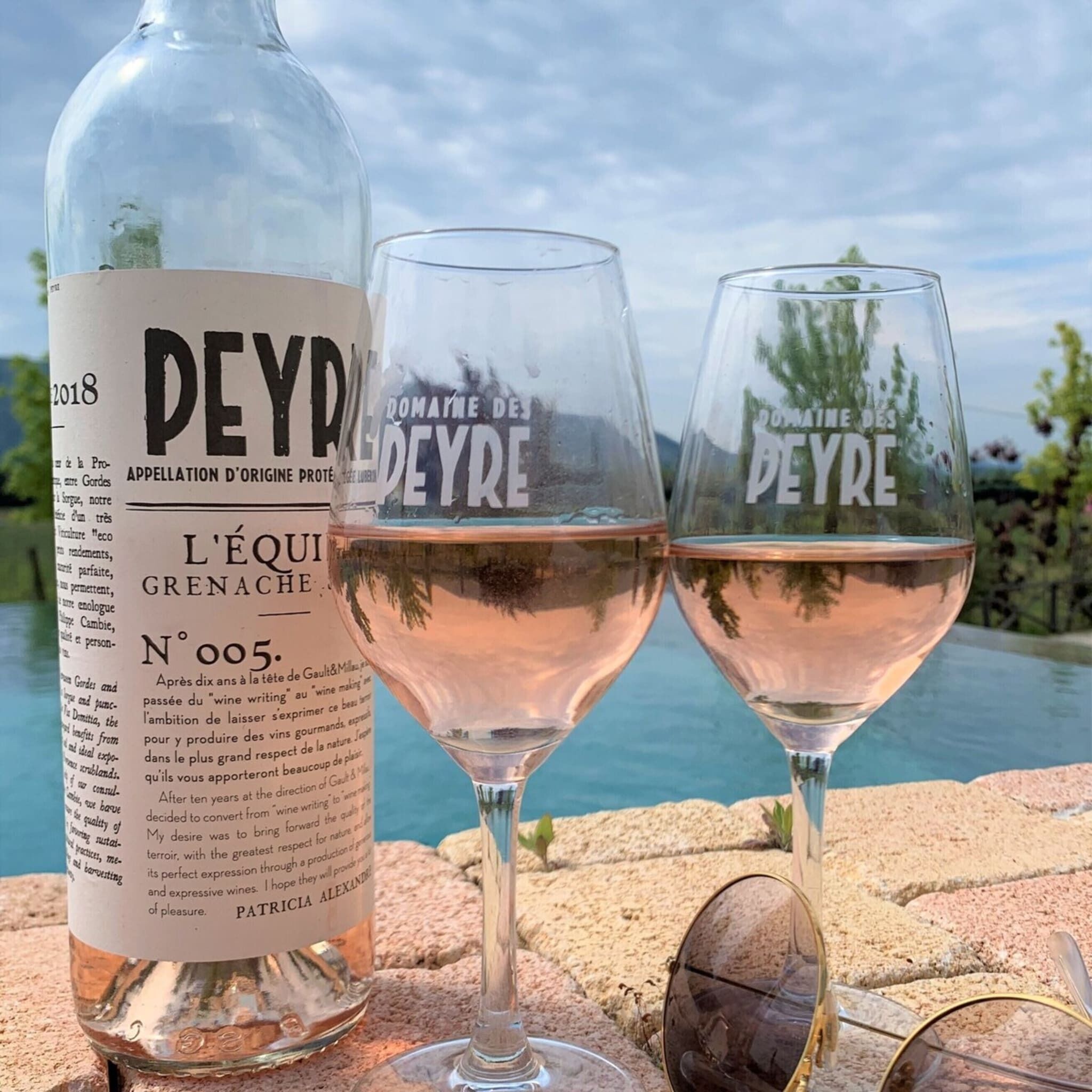 Une bouteille de vin Peyre et deux verres posés sur une surface en pierre, avec en arrière-plan un plan d’eau et de la végétation.