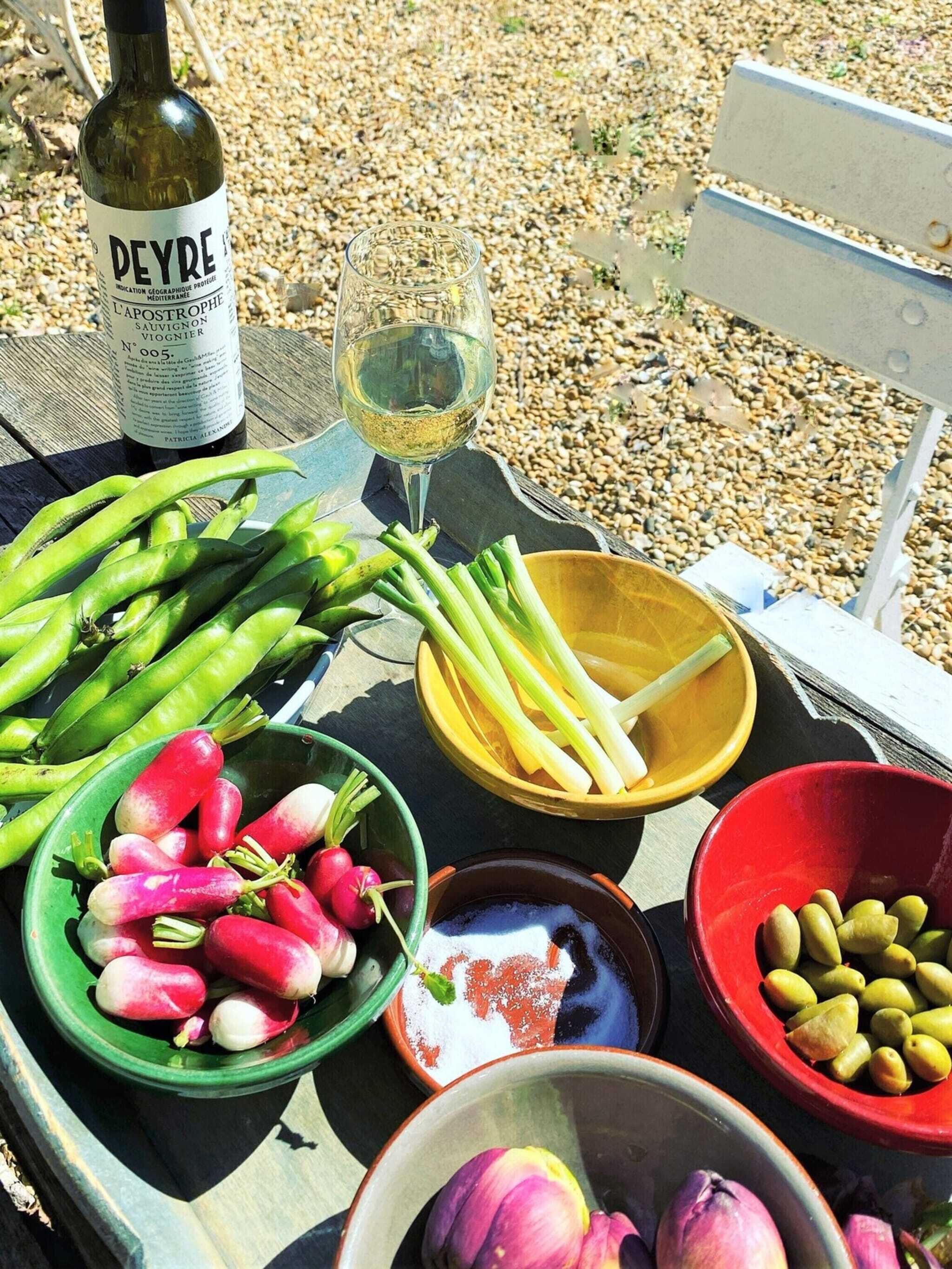 A table with a bottle of wine, a glass of white wine, and various vegetables including asparagus, radishes, celery, and olives.