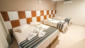 Ein Hotelzimmer mit drei Betten, jedes mit einer gestreiften Bettdecke. Das Zimmer verfügt über einen karierten Kopfteil und eine weiße Wand mit Lichtschalter und Klimaanlage.