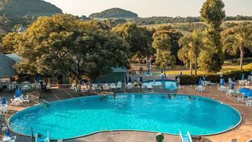 Ein Swimmingpool mit Rutsche, umgeben von Liegestühlen und Sonnenschirmen, vor einer Kulisse aus Bäumen und Hügeln.
