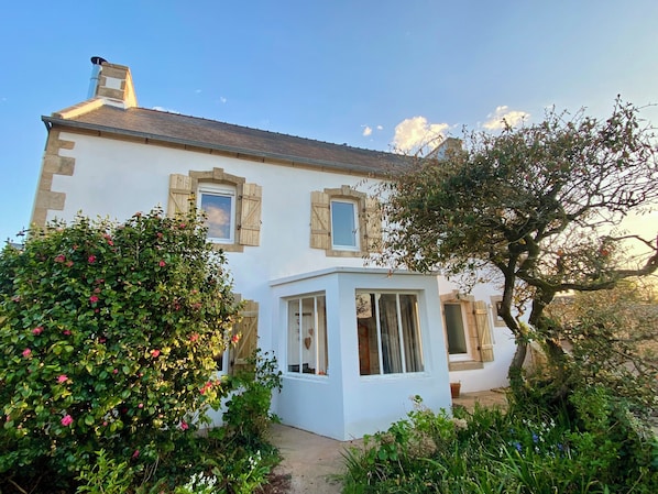 A two-story house with a white exterior, wooden shutters on the windows, and a small attached structure with glass doors. - Beachfront fisherman's cottage, family home style. (Plouhinec)