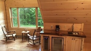 Interior de una cabaña con piso de madera, un comedor con sillas y mesa, una cocina con pileta y armarios, y ventanas amplias que dan a un bosque.