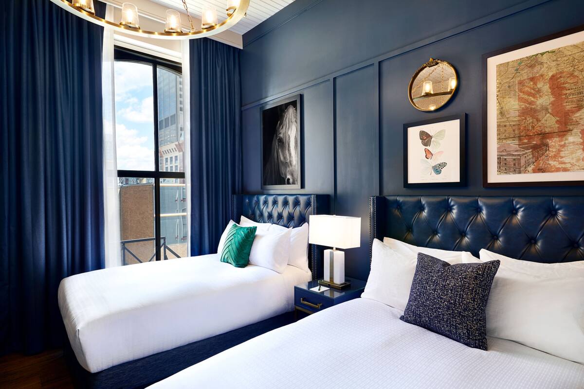 A hotel room with two beds, a large window with curtains, a wall-mounted mirror, and framed artwork.