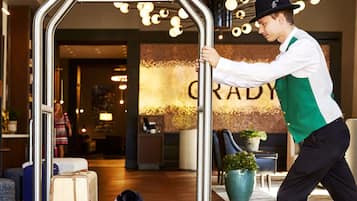Un hall d’hôtel avec un chariot à bagages, un chien noir allongé sur le chariot, et une personne qui pousse le chariot.