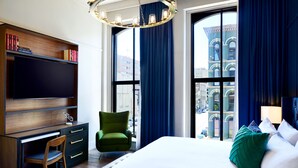 A modern hotel room with a large bed, a desk with a flat-screen TV, a green armchair, and a view of the city through large windows. - The Grady (Louisville)