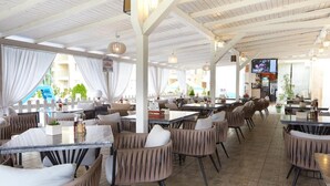A spacious outdoor dining area with multiple tables and chairs, a pool in the background, and a TV screen mounted on the wall. - Apart Hotel Vechna R (Nessebar)