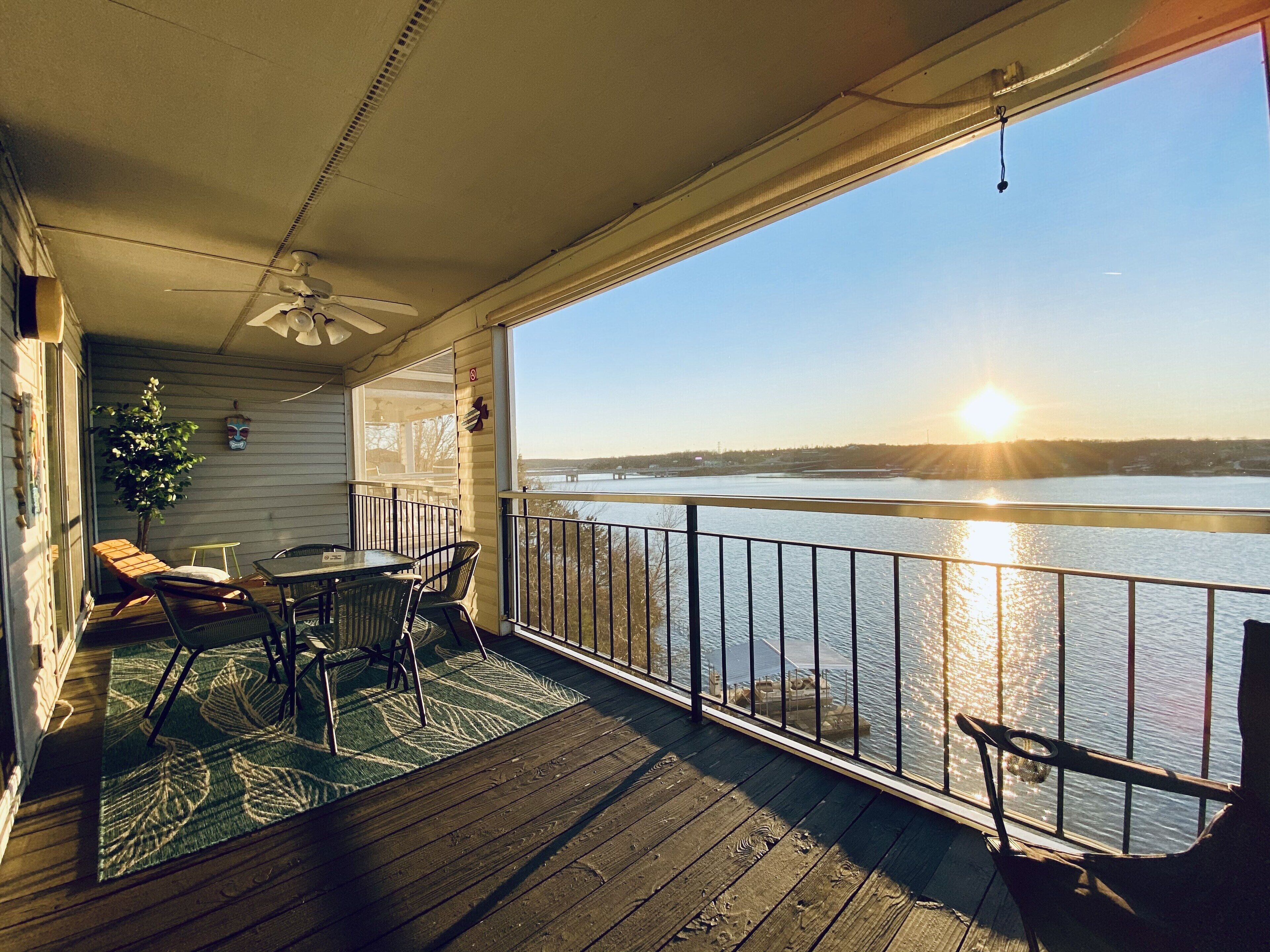 A wooden deck with a table and chairs, overlooking a body of water with a sunset.