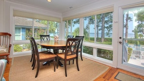 A dining area with a round table and chairs, large windows with a view of the sea and trees, and a wooden floor.