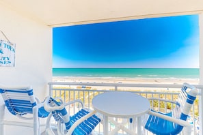 A balcony with a view of the beach, white and blue chairs, and a small table. - 5th floor oceanfront condo w/ shared pool, balcony, central AC, shared hot tub (North Myrtle Beach)
