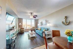 A spacious living room with a large window, a ceiling fan, a teal sofa, a wooden dining table, and a decorative anchor on the wall.