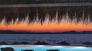 Una piscina infinita con sillones de descanso, vista al atardecer y techo de paja.